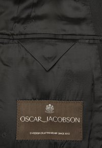 Wnętrze z czarnego satynowego materiału z brązową metką z wzorem jodełki, białym napisem: „OSCAR JACOBSON, SZWDZKIE RZEMIOSŁO OD 1903.”