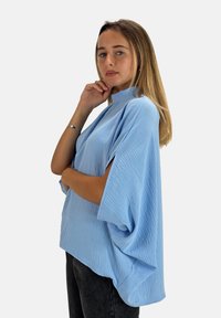 Maglietta oversized azzurro chiaro, strutturata, con maniche larghe e collo alto. Abbinata a jeans scuri, la modella posa con una mano sul mento.