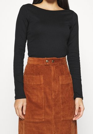 Mujer vistiendo un top negro de manga larga metido en una falda de pana color óxido de talle alto con bolsillos delanteros y detalles de botones.