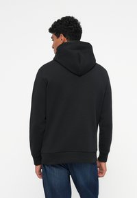 Sudadera negra con un bolsillo frontal tipo canguro, capucha con cordón y puños acanalados. Combinada con jeans azules, mostrando una textura suave y un ajuste casual.