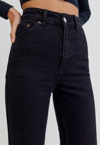 Jean noir taille haute en denim, doté de cinq poches, d'un bouton métallique et de rivets, avec une texture lisse et une décoloration minimale.