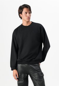 Homme portant un sweat-shirt noir oversized et un pantalon cargo en cuir noir, se tenant avec une main dans la poche, regardant sur le côté.
