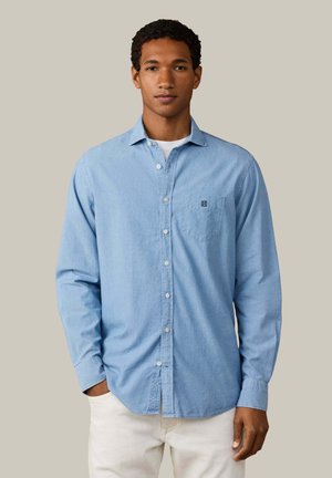 Jonge man draagt een lichtblauw overhemd over een wit ondershirt, staand met één hand in zijn zak tegen een effen achtergrond.