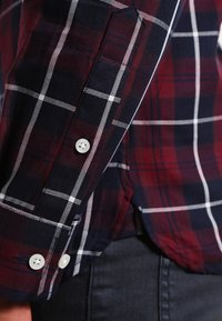 Primo piano di una manica di camicia scozzese rossa e blu navy con bottoni bianchi, indossata sopra un paio di jeans in denim blu scuro.