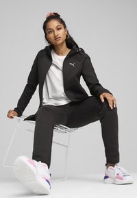 Chaqueta atlética negra con capucha y detalles de cremallera, combinada con una camiseta gráfica blanca y pantalones de chándal negros. El calzado presenta acentos en blanco, rosa y azul.