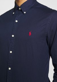 Mörkblå knapp-upp skjorta i bomull, med vita knappar, en liten röd broderad logotyp på bröstet och en klassisk krage.