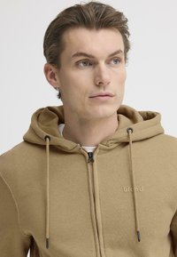 Brun hettejakke med glidelås i mykt stoff, med frontglidelås, snorjusteringer og en diskret logo på brystet.