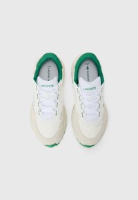 Witte stoffen sneakers met groene accenten, voorzien van een textuur bovenkant, platte veters en een dikke rubberen zool. "LACOSTE" logo weergegeven.