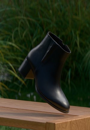 Bottine en cuir noir à talon carré flottant au-dessus d’un banc en bois, fond herbe verte.