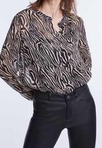 Gennemsigtig sort og beige zebra mønster bluse med knaplukning foran, kravefri design og afslappet pasform. Bæres med sorte slim-fit bukser.