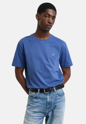 Camiseta de algodón azul con escote redondo y mangas cortas, que presenta un pequeño logo bordado en el pecho, combinada con jeans de mezclilla de lavado claro.