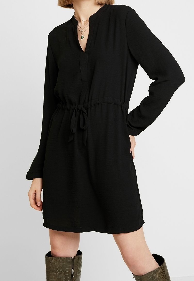 Femme portant une robe noire à manches longues avec une ceinture à la taille et des colliers superposés, associée à des bottes texturées montant jusqu'aux genoux.