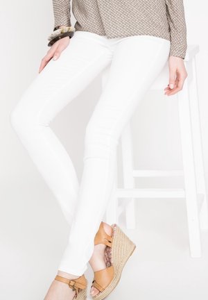 Białe, obcisłe dżinsy wykonane z elastycznego materiału, charakteryzujące się smukłym krojem i gładką fakturą. Noszone z beżowymi sandałami na koturnie.