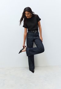 Femme en haut noir et jean large sombre ajustant une chaussure noire à bout pointu sur fond blanc uni.