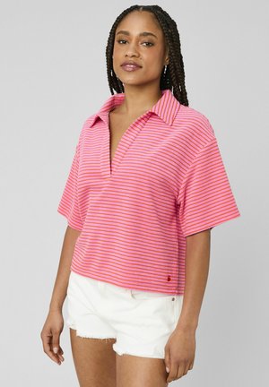 Femme aux longues tresses portant un t-shirt à manches courtes rayé rose et orange et un short en denim blanc, debout devant un fond gris.