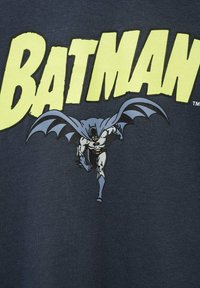 Mornarica majica s velikim žutim natpisom "BATMAN" i slikom Batmana u plavoj i sivoj boji, prikazanog u dinamičnoj poziciji trčanja s crnim akcentima.