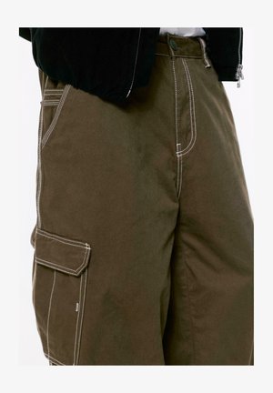 Nahaufnahme von dunkelolivfarbenen Cargo-Hosen mit weißen Kontrastnähten und einer seitlichen Taschenklappe, getragen mit einer schwarzen Jacke und einem weißen Hemd.