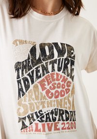 Camiseta de algodón blanca con un diseño tipográfico estilizado multicolor que incluye frases en negro, rojo y dorado en el frente.
