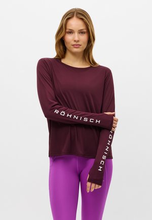 Vrouw staat met armen over elkaar, draagt een donkerrode top met lange mouwen en witte tekst op de mouwen en felpaarse leggings.