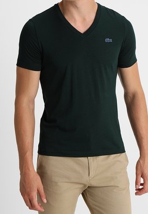 Mann trägt ein eng anliegendes dunkelgrünes V-Ausschnitt-T-Shirt mit kleinem gesticktem Logo auf der Brust und beigen Hosen, steht mit entspannten Armen.