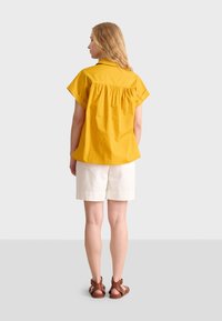 Camicia gialla senape a maniche corte con dettaglio arricciato sul retro, abbinata a pantaloni corti bianchi. Modella che indossa sandali marroni, in piedi davanti a uno sfondo neutro.