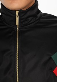 Svart zip-up jacka med hög krage, guldzipper och textilstruktur; har röda och gröna detaljer på vänster sida.