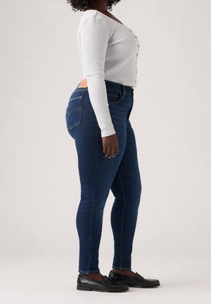 Kobieta w ciemnoniebieskich obcisłych dżinsach, białym prążkowanym topie z długim rękawem i guzikami oraz czarnych mokasynach, stojąca bokiem na jednolitym tle.