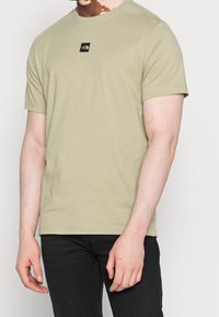 Ljus olivgrön t-shirt med korta ärmar i bomull, med en liten svart logotyp på bröstet och rund halsringning.