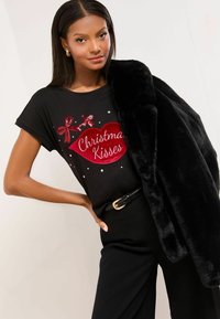 Melna T-kreklā ar sarkaniem sequīnu lūpām un uzrakstu "Ziemassvētku skūpsti", apvienota ar melnu mākslīgās kažokādas mēteļu un melnu jostu.