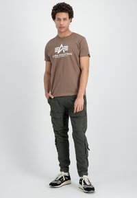 Camiseta de algodón marrón con el logo blanco "Alpha Industries", combinada con pantalones cargo verde oliva que tienen bolsillos laterales y puños ajustables.
