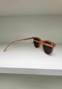 Bruine plastic zonnebril met ronde zwarte lenzen en licht gebogen pootjes, met "Komono" branding aan de zijkant.