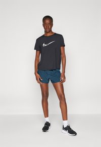 Svart Nike-t-skjorte med hvit logo, kombinert med blå shorts. Modellen har på seg svarte treningssko og hvite sokker. Minimalistisk design, lett stoff.