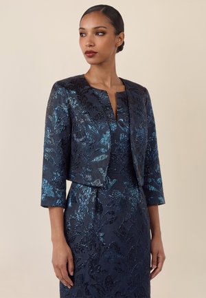 Frau trägt eine marineblaue, strukturierte Blumenjacke mit dreiviertel Ärmeln über einem passenden Kleid, steht vor einem beigen Hintergrund.