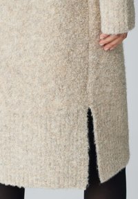 Beiger Wollmantel mit strukturiertem Finish, seitlichem Schlitz und langen Ärmeln. Der Stoff wirkt weich und leicht fuzzy.