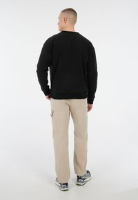 Zwart sweatshirt met ronde hals, gedragen over beige cargobroek, met instap schoenen met een gestructureerde zool en een patroon.