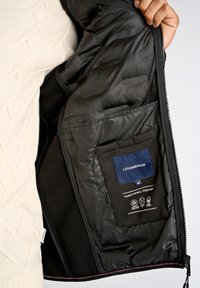 Svart inre foder av en pufferjacka, med ett etikett märkt "LINDBERGH," och en ficka som visar skötselsymboler och designdetaljer.