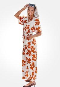 Lang, crème-kleurig jurkje met grote roestkleurige bloemenpatronen, korte mouwen, elastische taille en zakken. In combinatie met bruine, puntige hakken.