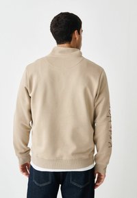 Beige sweatshirt med høj krave, ribkant ved ærmer og bund. Har brun tekst langs ærmet. Blød tekstur, afslappet pasform.