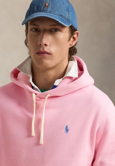 Jaunas vyras dėvi mėlyną džinsinę kepurę su siuvinėtu arkliu, sluoksniuotus rožinius megztinius su gobtuvu ir baltą marškinių apykaklę, neutrali fonas.