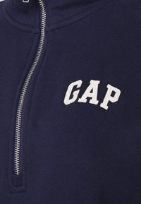 Gros plan d’un vêtement bleu marine zippé avec logo GAP blanc brodé sur la poitrine et une fermeture éclair argentée visible.