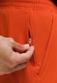 Eine Hand hält den Reißverschluss einer orangen Kleidungsstücktasche, zieht ihn teilweise auf und zeigt die Struktur des Stoffes und die Nähte.