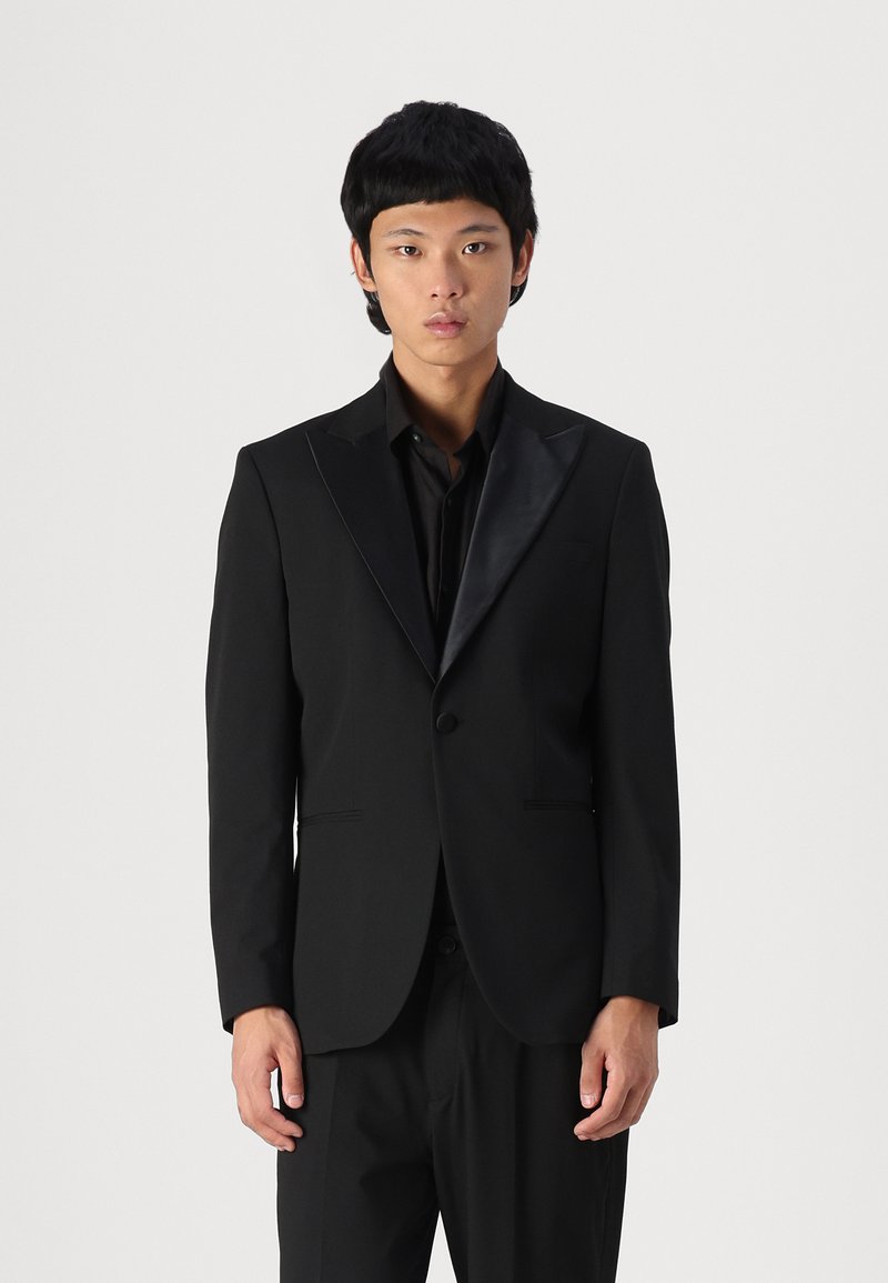 Black tuxedo jacket with a glossy satin peak lapel, single button closure, and tailored fit. Worn with a matching black shirt and trousers.