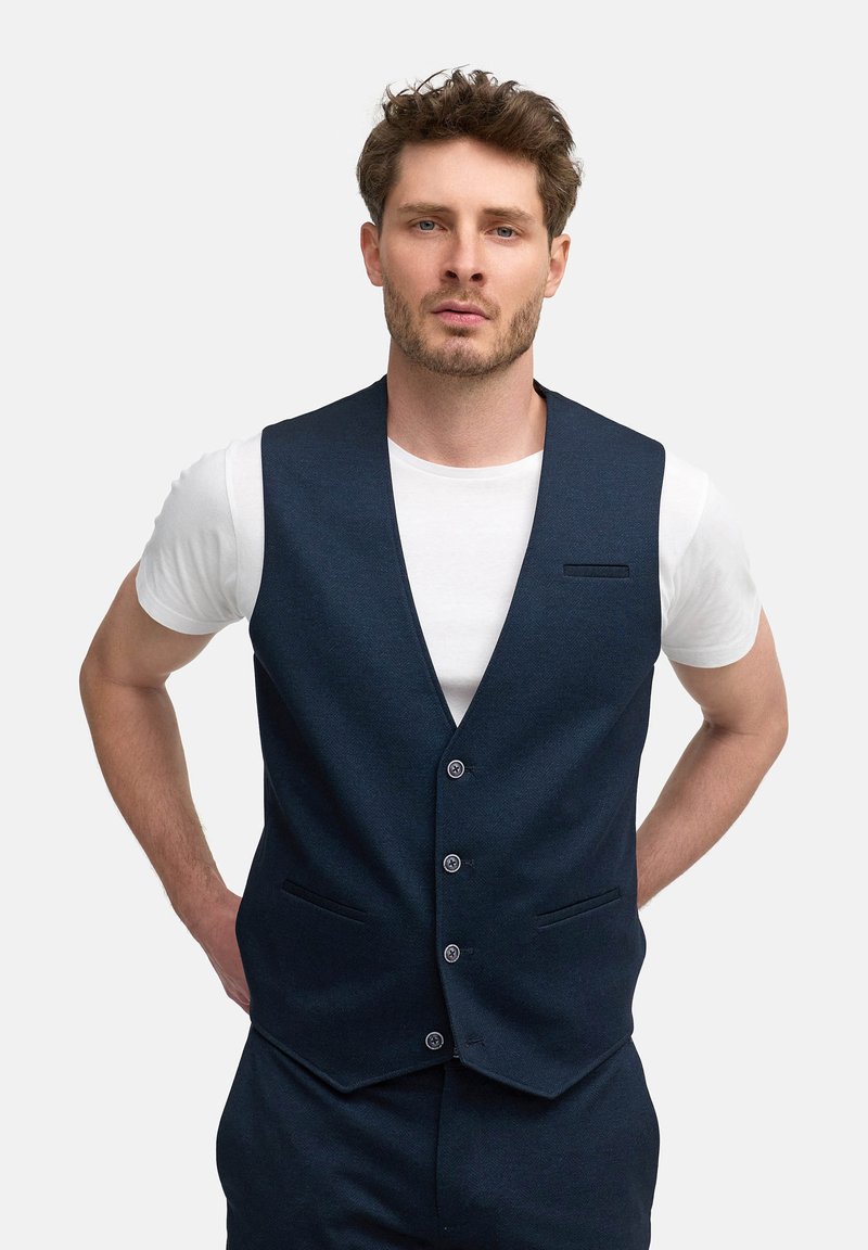 Chaleco de color azul marino oscuro con una tela texturizada, que presenta un escote en V, cinco botones y dos bolsillos laterales. Se lleva sobre una camiseta blanca.
