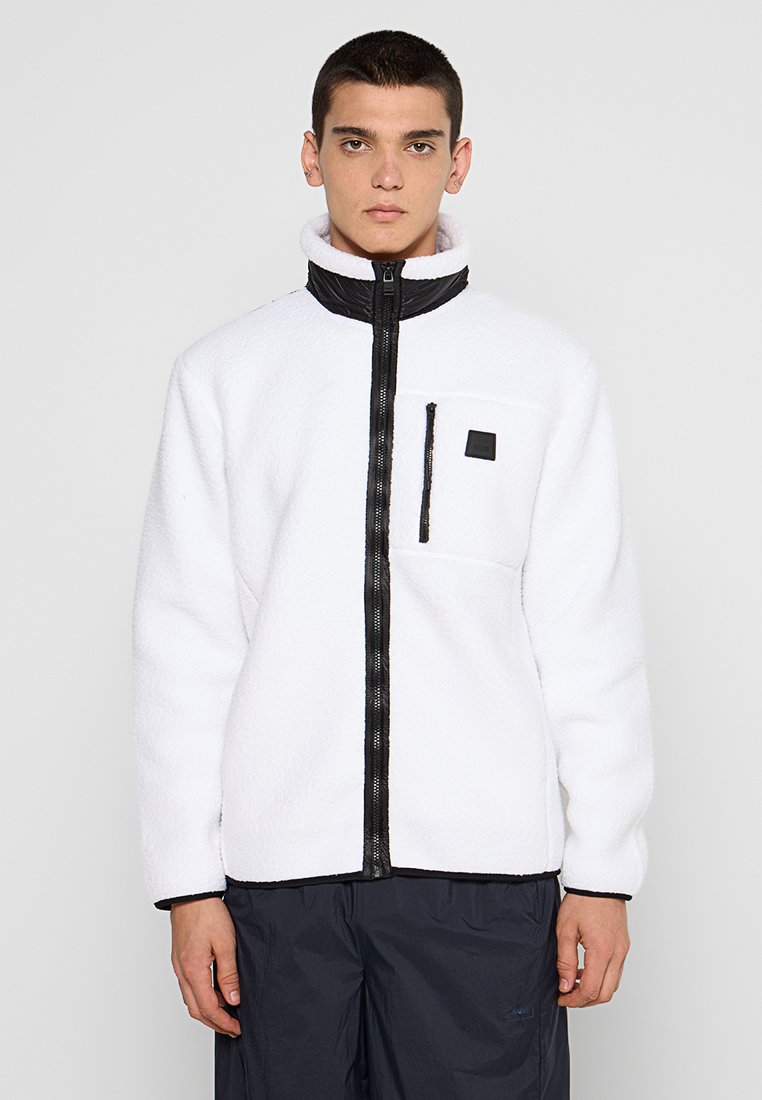Jeune homme portant une veste en polaire blanche avec une fermeture éclair noire et une poche sur la poitrine, se tenant devant un fond gris uni.