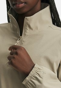 Beige pullover kõrge kraega ja poole zip-sulguriga; omab elastseid mansette ja tekstuurset kangast koos diskreetse logo aktsendiga.