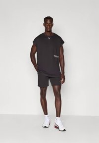 Mies seisoo yksivärisen taustan edessä, yllään musta hihaton Puma-paita, mustat shortsit, valkoiset Puma-sukat ja valkoiset urheilukengät.