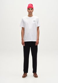 Tricou alb din bumbac cu imprimeu negru pe partea stângă a pieptului, purtat cu pantaloni negri, pantofi maro și o căciulă tricotată roz aprins.