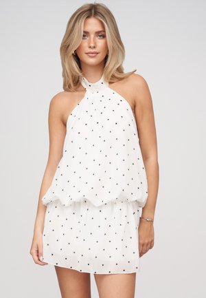 Donna bionda che indossa un vestito bianco senza maniche con pois neri, in piedi davanti a uno sfondo chiaro e uniforme.
