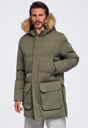 Homme portant un parka d'hiver vert olive avec capuche doublée de fourrure, poches avant et fermeture éclair latérale, debout devant un fond uni.