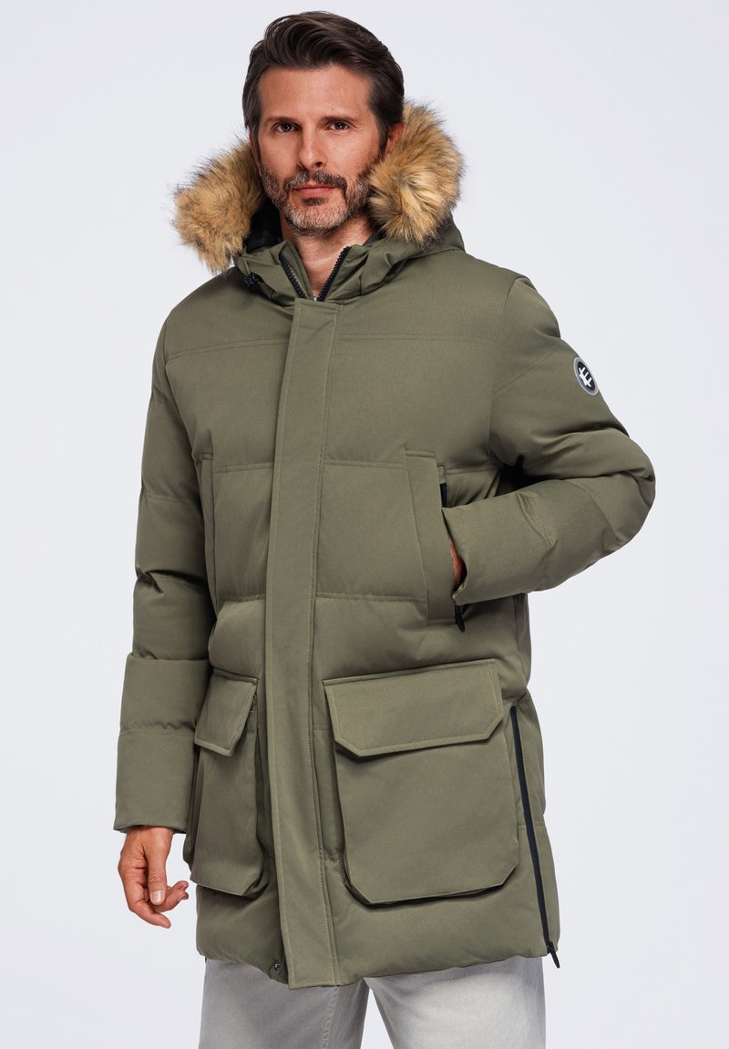 Homme portant un parka d'hiver vert olive avec capuche doublée de fourrure, poches avant et fermeture éclair latérale, debout devant un fond uni.